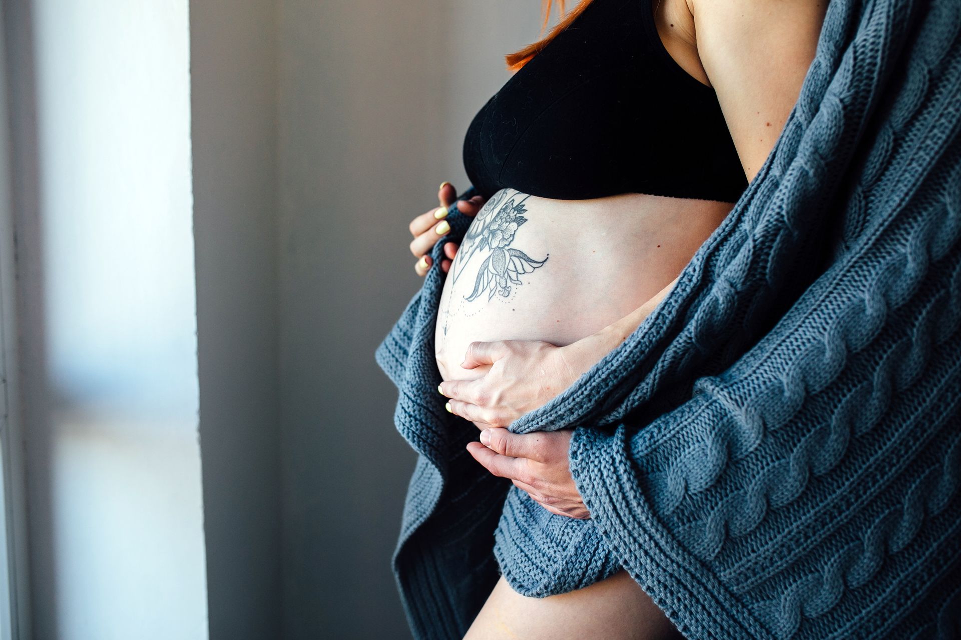 Pregnant woman in a knit cardigan cradling her belly, which has a floral tattoo, raising the question of getting a tattoo while pregnant or breastfeeding