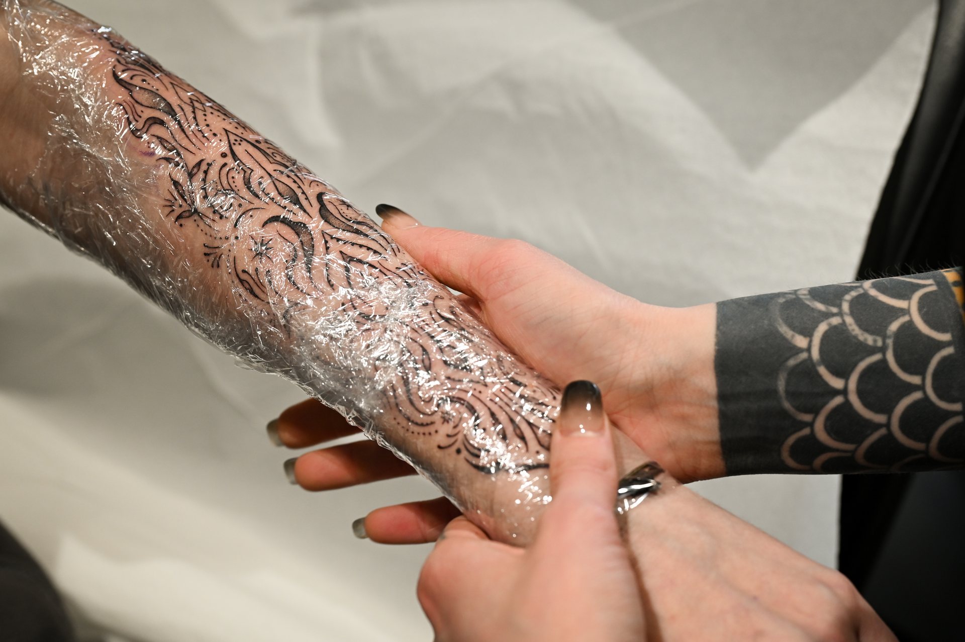Artist wrapping a freshly inked, intricate black-and-grey forearm tattoo in protective plastic film during the tattoo healing process