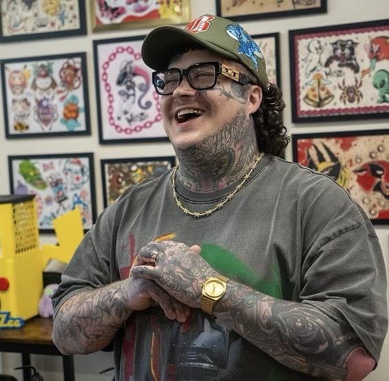 Matt Zee, tattoo artist at Enigma Tattoos, smiling in the studio with framed flash art on the wall behind him.